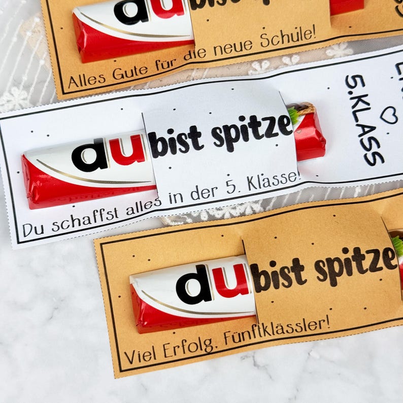 May include: Three chocolate bars wrapped in red and white packaging, each secured with a paper sleeve. The sleeves feature black text including 'du bist spitze', 'Alles Gute f&uuml;r die neue Schule', '5. KLASS', and 'Du schaffst alles in der 5. Klasse'.