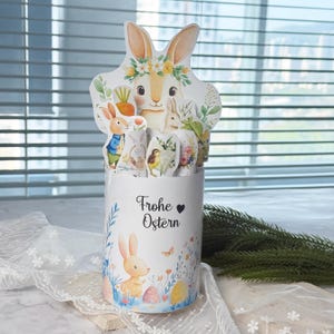 May include: A white cylindrical Easter-themed container with a bunny and floral design. The container has a bunny-shaped topper and includes several small, decorative Easter-themed cutouts. The text "Frohe Ostern" is printed on the front.