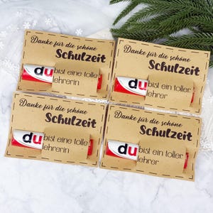 May include: Four rectangular kraft paper cards with black text reading Danke für die schöne Schulzeit. Each card features a red and white Duplo chocolate bar attached with a paper band printed with bist eine tolle lehrerin or bist ein toller lehrer.