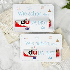 Puede incluir: Dos tarjetas rectangulares de papel blanco con el texto en alemán "Wie schön, dass du DA BIST" en azul y negro. Cada tarjeta sostiene una barra de chocolate roja y blanca con la palabra "du" visible. Pequeños iconos escolares decoran las tarjetas.