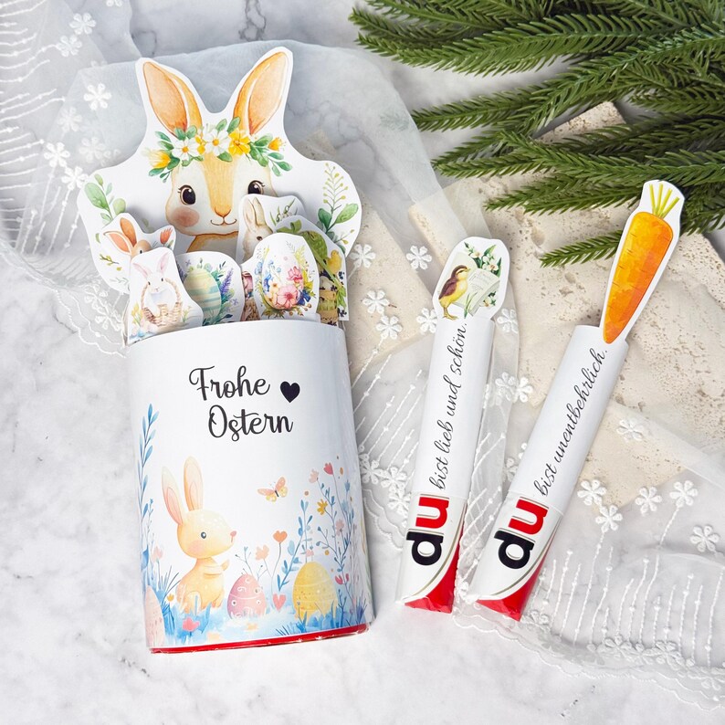 May include: A white Easter-themed container with a bunny and floral design, holding bunny and egg cutouts. The container has the text "Frohe Ostern". Two white candy sticks with carrot and bird designs are also present.