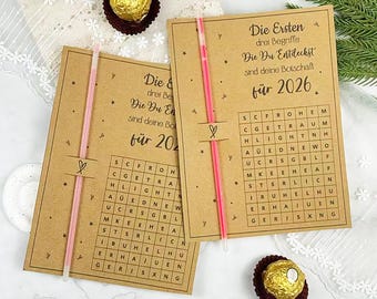 Könnte beinhalten: Zwei Karten aus Kraftpapier mit rosa Akzenten und einem Wortsuchrätsel. Die Karten haben deutschen Text, darunter "für 2026". Ein rosa Strohhalm und eine Praline sind ebenfalls sichtbar. Die Karten sind mit einem Herzsymbol verziert.