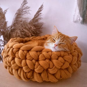 Pode incluir: Uma cama redonda para animais de estimação em malha grossa de cor amarelo-mostarda quente. A cama apresenta uma textura de fio trançado espesso com uma abertura circular. Um gato malhado laranja com marcas brancas repousa no seu interior.