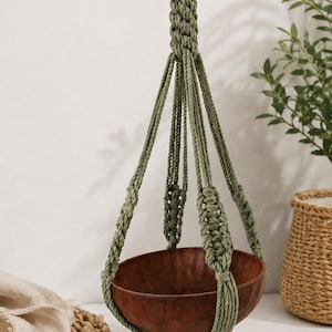 Puede incluir: Un soporte colgante para plantas que presenta un cuenco de madera de color marrón oscuro suspendido por un cordón de macramé de color verde salvia. El cordón está intrincadamente anudado con un lazo central en la parte superior para colgar.