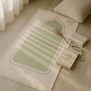 May include: A prayer rug with a light green arched design on a cream background. A cream-colored pillow, a book, prayer beads, and folded cloths are next to the rug. Dried flowers are on the rug.