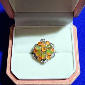 May include: A silver-toned ring featuring a cluster of oval-shaped, iridescent orange opals arranged in a floral pattern, accented with small, clear, round-cut stones along the scalloped metal edges, presented inside a light pink jewellery box.