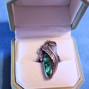 May include: A silver ring featuring an elongated, iridescent green abalone inlay set within a metal leaf-shaped frame. The ring is displayed inside a light-colored, octagonal jewelry box with a soft, white interior lining and a hinged lid.