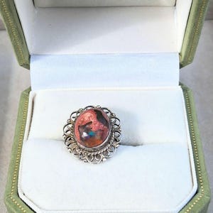 May include: A silver ring featuring an oval-shaped stone with a mottled surface of reddish-brown, black, and iridescent blue flecks. The stone is set within a decorative, scalloped silver frame with a twisted wire border detail.