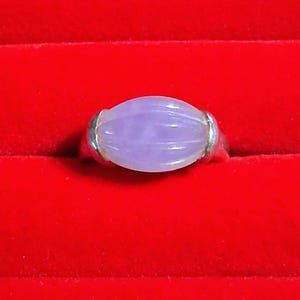 May include: A ring featuring a smooth, oval-shaped, light purple stone with vertical ribbed detailing. The stone is set in a polished silver-toned metal band that curves around the sides of the gemstone, securing it firmly in place.