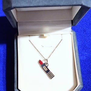 May include: A silver-toned necklace featuring a lipstick-shaped pendant with a red tip and a rectangular base. The pendant hangs from a delicate chain, presented inside a dark gray, octagonal-shaped jewelry box with a cream-colored interior lining.