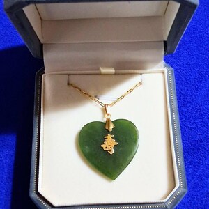 May include: A heart-shaped dark green jade pendant suspended from a gold chain. The pendant features a central gold-colored Chinese character for longevity. The necklace is presented inside a dark gray, octagonal-shaped jewelry box with a white interior.