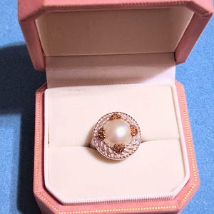 May include: A ring featuring a central white pearl surrounded by a textured silver-tone metal border with small, embedded orange-brown stones. The ring is presented inside an open, octagonal, light pink velvet jewelry box with a cream interior.