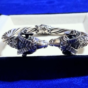 May include: A silver cuff bracelet featuring two detailed ram heads facing each other at the opening. The band has a twisted rope design with intricate filigree metalwork accents near the ram heads, showcasing a polished and oxidized finish.