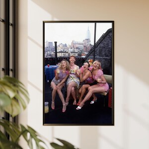 K&ouml;nnte beinhalten: Gerahmtes Foto mit f&uuml;nf lachenden Frauen, die Getr&auml;nke auf einem Dach mit Skyline im Hintergrund halten. Die Frauen tragen Sommerkleidung, darunter Kleider und Shorts. Das Foto wird an einer beigefarbenen Wand gezeigt.