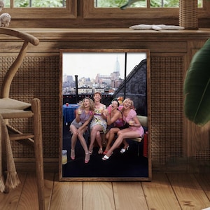 K&ouml;nnte beinhalten: Gerahmtes Foto mit f&uuml;nf lachenden Frauen, die Getr&auml;nke auf einem Dach mit Skyline im Hintergrund halten. Die Frauen tragen Sommerkleidung, der Rahmen ist aus dunklem Holz.