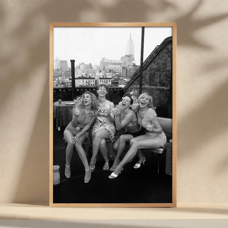 K&ouml;nnte beinhalten: Schwarz-wei&szlig; gerahmtes Foto von f&uuml;nf lachenden Frauen auf einem Dach mit Skyline im Hintergrund. Die Frauen tragen Kleider und Abs&auml;tze und halten Getr&auml;nke. Das Empire State Building ist sichtbar.