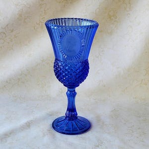 May include: A cobalt blue glass goblet featuring a flared rim with vertical fluting, a central cameo profile portrait surrounded by a scalloped border, and a diamond-patterned bowl. The stem is faceted, leading to a circular, rayed base.
