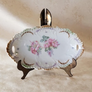 May include: Oval porcelain serving dish with a scalloped edge, featuring a hand-painted floral design in pink, purple, and green. The dish has small handles and is supported by a metal stand. A decorative piece for serving.