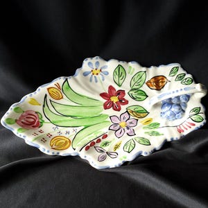 May include: A white ceramic leaf-shaped serving dish with a scalloped edge, hand-painted with colorful flowers, green leaves, and fruit. The dish has a blue rim and a small handle. The background is a dark fabric.
