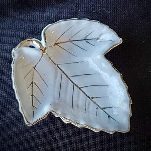 May include: A white, leaf-shaped dish with gold trim and gold veining details. The dish has a glossy finish and is likely made of ceramic or porcelain. The design is elegant and would be suitable for serving small items.