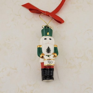 May include: A ceramic Christmas ornament shaped like a toy soldier drummer. The ornament is white with green and gold accents, black boots, and red pants. A red ribbon is attached for hanging.
