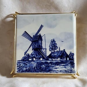 May include: A square ceramic tile featuring a blue and white illustration of a traditional windmill and a small cottage. The tile is held within a gold-toned metal frame with curved corner accents, creating a decorative display piece.
