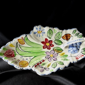 May include: A white ceramic serving dish shaped like a leaf with a scalloped edge. The surface features hand-painted floral motifs in red, purple, yellow, and green, with a three-dimensional cluster of blue grapes near the integrated handle.