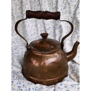 May include: A copper tea kettle with a rounded body, a short spout, and a removable lid featuring a small knob. The kettle has a curved metal handle topped with a dark brown wooden grip, showing a weathered patina across the metallic surface.