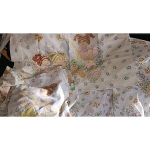 May include: A white fabric sheet set featuring a repeating pattern of Cabbage Patch Kids characters, including dolls with red, yellow, and brown hair, alongside colorful butterflies, flowers, and rainbows. The fabric has a soft, printed design.