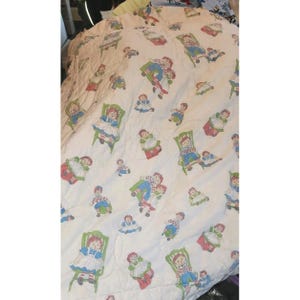 Peut inclure: Couette blanche avec un motif répété d'une poupée en robe bleue, d'un garçon en salopette et d'une chaise verte. La couette a un motif en losange et est probablement en coton.
