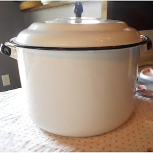 May include: A large, white enamel pot with a lid and black trim. The pot has two side handles and a black knob on the lid. The pot is sitting on a pink and white patterned surface.