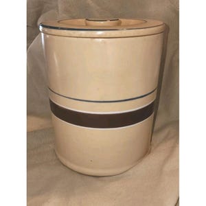 Op de afbeelding: Een beige keramische pot met een deksel. De pot heeft een donkerbruine band met een witte streep en een dunne blauwe streep. Het deksel heeft een klein handvat en een dunne blauwe streep. De pot is cilindrisch.