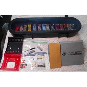 Könnte beinhalten: Ein schwarzer Koffer mit transparentem Deckel enthält mehrere Miniatur-Skateboards. Weitere Artikel sind eine rot-schwarze Aufbewahrungseinheit, eine graue Box mit dem Text "Tony Hawk Foundation" und verschiedene Kleinteile.