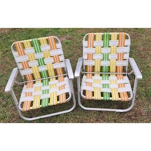 Puede incluir: Dos sillas de jardín plegables vintage con asientos y respaldos tejidos. Los asientos presentan un patrón colorido de tiras tejidas naranjas, amarillas, verdes y blancas. Los marcos son de metal plateado, y las sillas están sobre hierba.