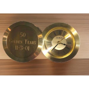 May include: A gold-colored Danbury Clock Company quartz clock with a hinged lid. The lid is engraved with "50 Golden Years 11-3-01." The clock face has Roman numerals and black hands. The clock is open, revealing the time.
