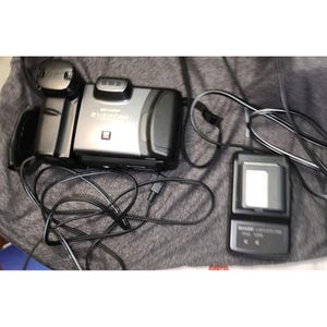 May include: A grey Sharp Viewcam camcorder with a black battery pack attached, accompanied by a separate black battery charger unit. Both devices are connected by black power cords, and the camcorder features the Sharp Viewcam logo on its front.