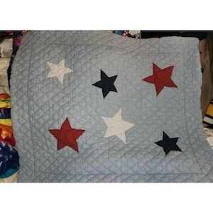 May include: A light blue quilted blanket featuring a pattern of five appliqued stars. The stars are arranged in a scattered layout and alternate between solid white, dark navy blue, and deep red colors across the textured fabric surface.