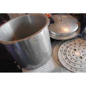 Puede incluir: Una olla a presión grande, abierta, de color plateado, con tapa e inserto perforado. La olla está hecha de metal y parece usarse para cocinar o enlatar. La tapa tiene un asa y el inserto tiene muchos agujeros pequeños.