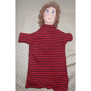 May include: A handmade puppet with a painted face, brown curly hair, and blue eyes. The puppet wears a red and black striped shirt. The puppet's arms are outstretched, and the background is a dotted pattern.