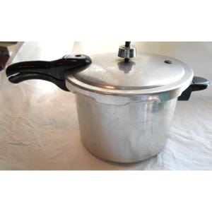 Könnte beinhalten: Ein silberner Schnellkochtopf mit schwarzem Griff und Deckel. Der Kocher hat eine glänzende Metalloberfläche und ein Überdruckventil oben. Der Griff ist aus schwarzem Kunststoff. Der Kocher ist für schnelles Kochen von Speisen konzipiert.