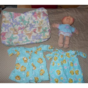 May include: A baby doll with a blue top and diaper, a matching tote bag with teddy bear pattern, and two blue duck-themed baby outfits. The outfits have yellow duck graphics on a light blue background.
