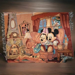 Puede incluir: Una manta de tapiz tejida que presenta una escena acogedora con Mickey y Minnie Mouse acurrucados en un sofá. La manta tiene una chimenea, una ventana con un muñeco de nieve y Pluto. La paleta de colores incluye tonos cálidos.