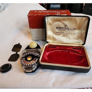 May include: A vintage Norwood Director exposure meter with accessories, including a case and a box. The meter has a black and silver face with a dial and a light sensor. The case is black with a red velvet interior, and the box is red and cream.