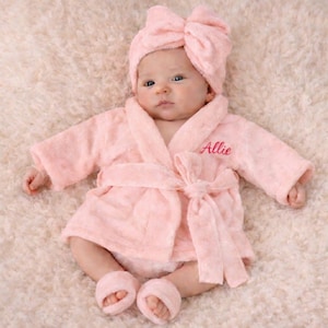 May include: A baby wearing a light pink terry cloth robe, matching bow-shaped headband, and booties. The robe is tied with a belt and has the name "Allie" embroidered on the chest. The baby is lying on a soft, light-colored surface.
