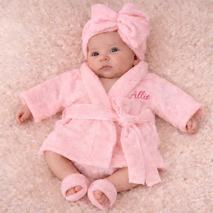 May include: A baby wearing a light pink terry cloth robe set, including a matching bow headband, robe, and booties. The name "Allie" is embroidered on the robe in a matching pink thread. The baby is lying on a light pink, fluffy surface.