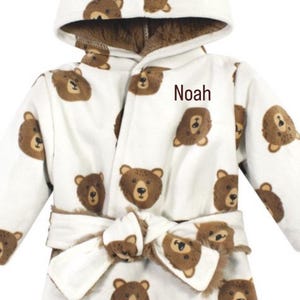 May include: White hooded baby bathrobe with a repeating pattern of brown bear faces. The robe has a matching belt and the name "Noah" is printed on the front. The interior of the hood and cuffs are a darker brown.