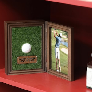 May include: A two-panel wooden frame with a golf theme. One side features a golf ball on artificial turf and a plaque with text. The other side displays a photo of a golfer. The frame is brown and sits on a red surface.