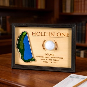 May include: A framed wooden display featuring a white golf ball centered between engraved text reading HOLE IN ONE, NAME, DOMINION VALLEY COUNTRY CLUB, HOLE 9 - 165 YARDS, and APRIL 9TH 2026, with a green and blue golf course map cutout on the left.