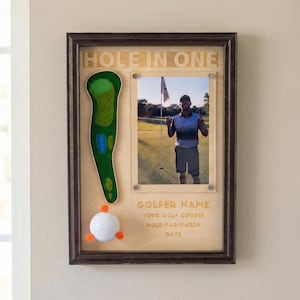 May include: Wooden framed golf-themed display with the words "HOLE IN ONE" at the top. It features a photo of a golfer, a green felt golf course design, and a golf ball with orange accents. The display includes space for golfer details.