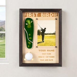 May include: A dark wood framed display case featuring the text FIRST BIRDIE at the top. It includes a green golf course map cutout, a white golf ball on a stand, and a rectangular photo frame with a picture of a golfer. Text includes YOUR NAME, GOLF CLUB, HOLE 5 - 167 YARDS, and YEAR AND DATE.